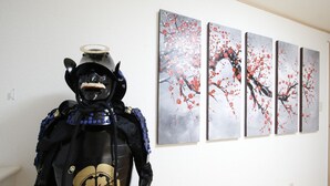 En skyltdocka klädd i en svart och guldfärgad samurajdräkt står i ett konstgalleri. På väggen hänger fyra målningar som föreställer körsbärsblommor.