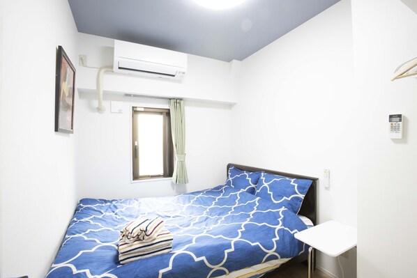 A small, single-bed room with a blue patterned bedspread, a bedside table, a wall-mounted air conditioner, a window with a curtain, and a framed picture on the wall. - Suginami Koenji Apartment (Tokyo)