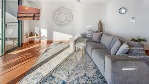 A modern living room with a sofa, a patterned rug, a wall clock, and a decorative wall piece. - Oceanfront Views at Moffat Beach + Rooftop Terrace (Moffat Beach)