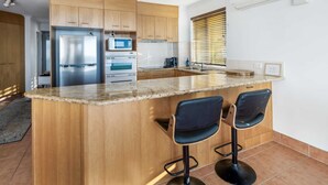 A kitchen with wooden cabinets, a stainless steel refrigerator, a microwave, and a sink. There are two bar stools at the island. - Oceanfront Views at Moffat Beach + Rooftop Terrace (Moffat Beach)