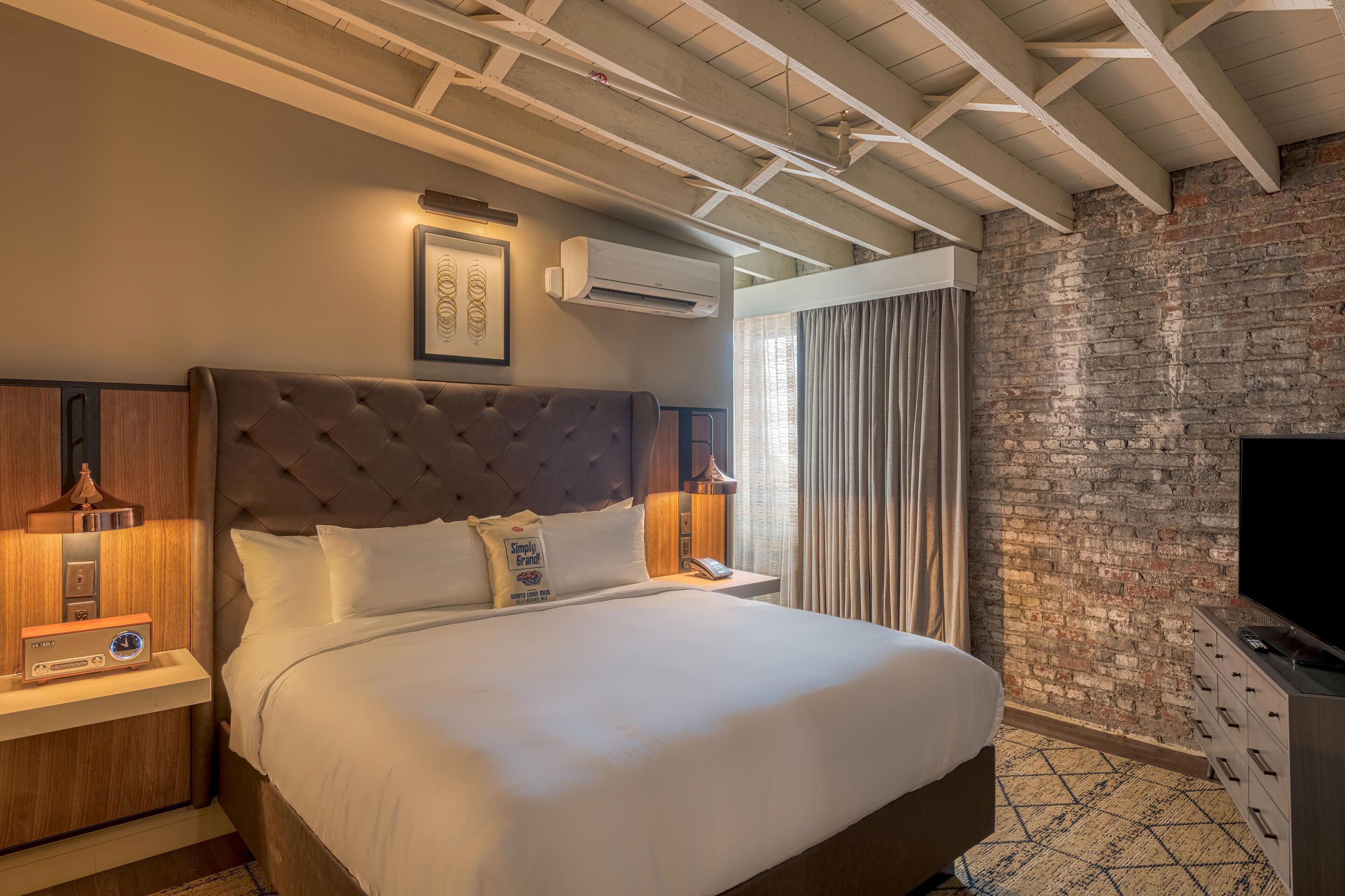 A hotel room with a large bed, a flat-screen TV, a brick wall, and a ceiling with exposed beams.