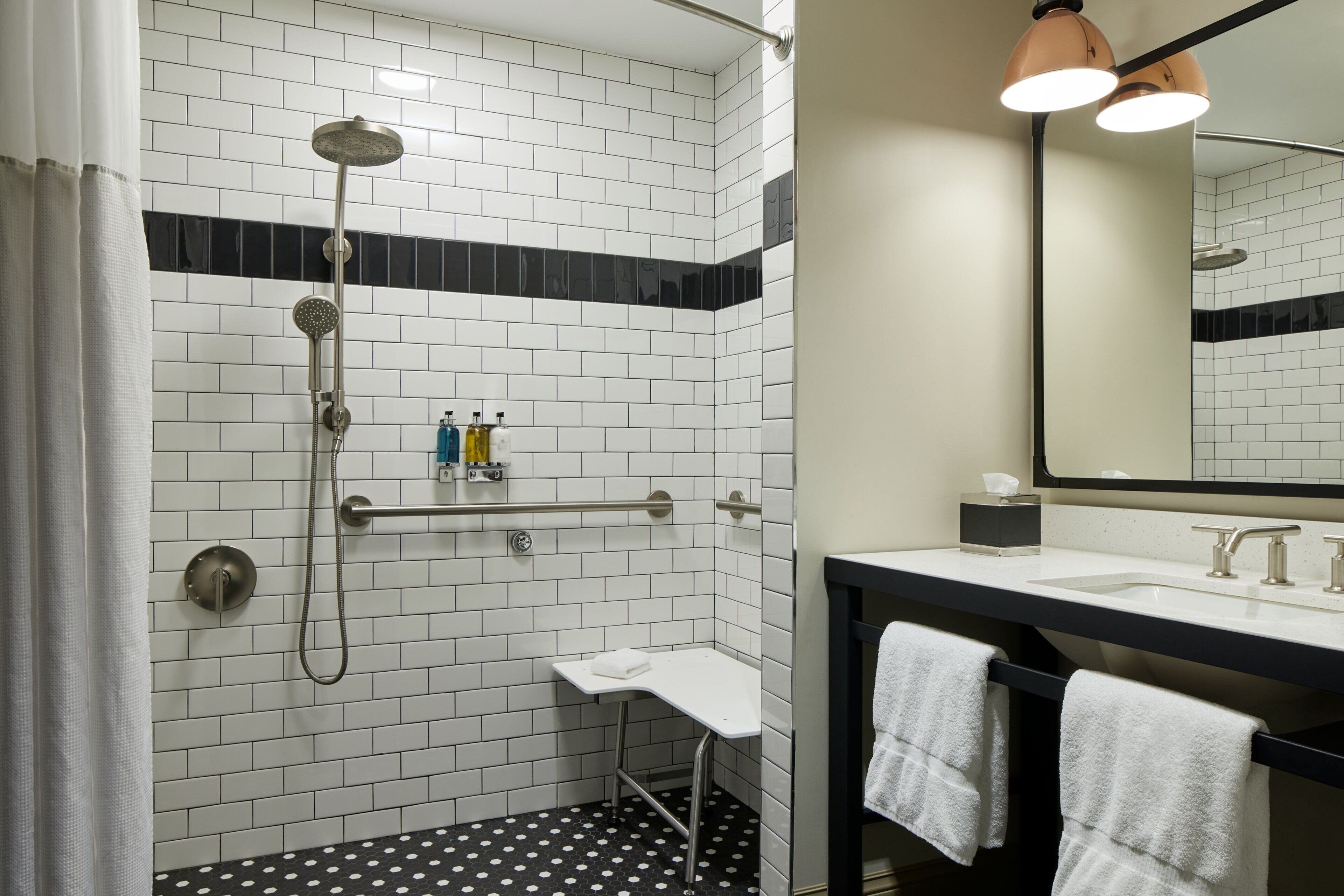 A modern bathroom with white and black tiled walls, a shower with a handheld showerhead, a white sink with two faucets, and a mirror above the sink.