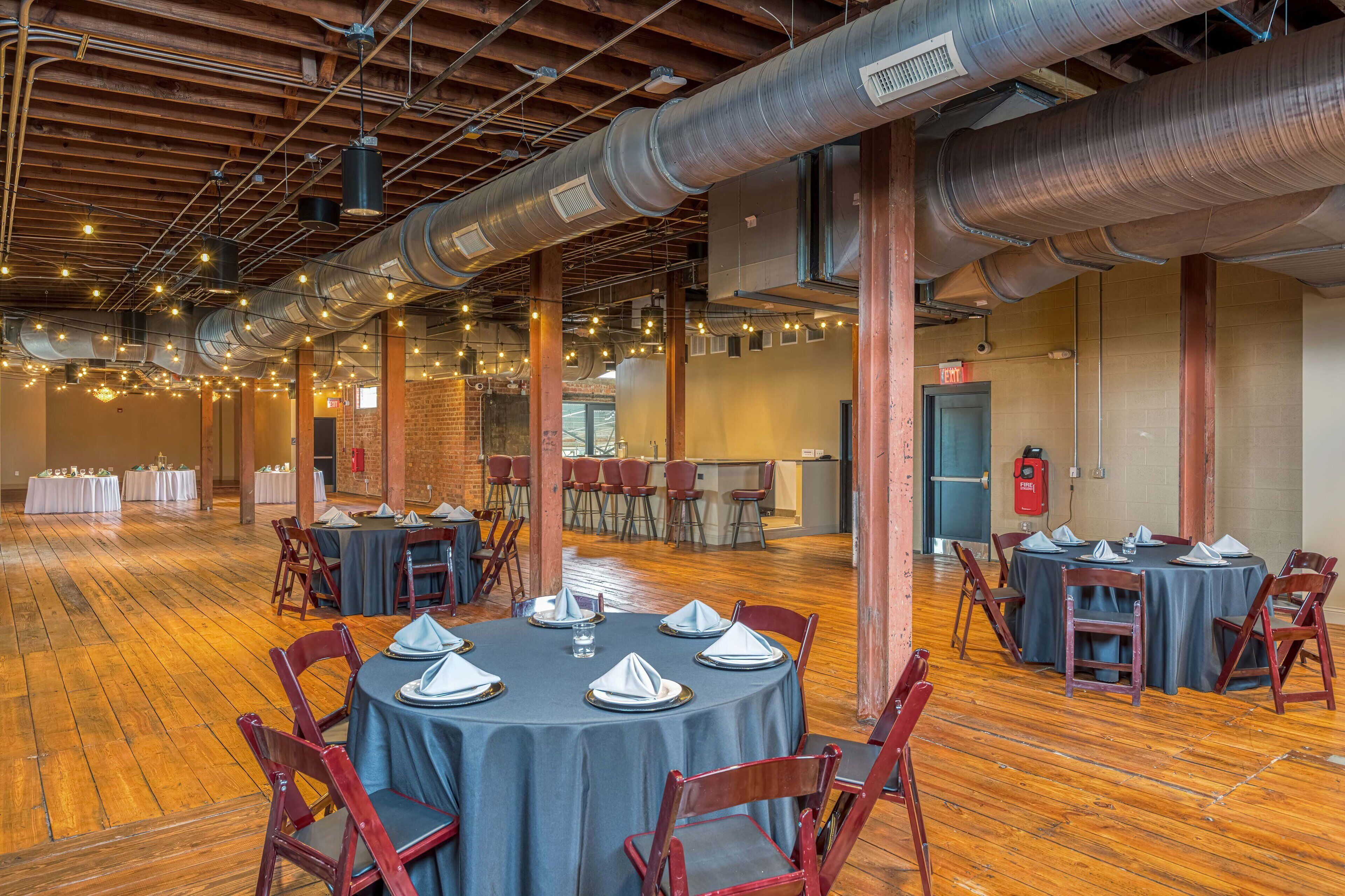 A spacious hall with round tables set for dining, exposed ductwork, and a brick wall in the background.