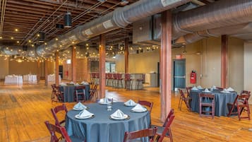 A spacious hall with round tables set for dining, exposed ductwork, and a brick wall in the background.