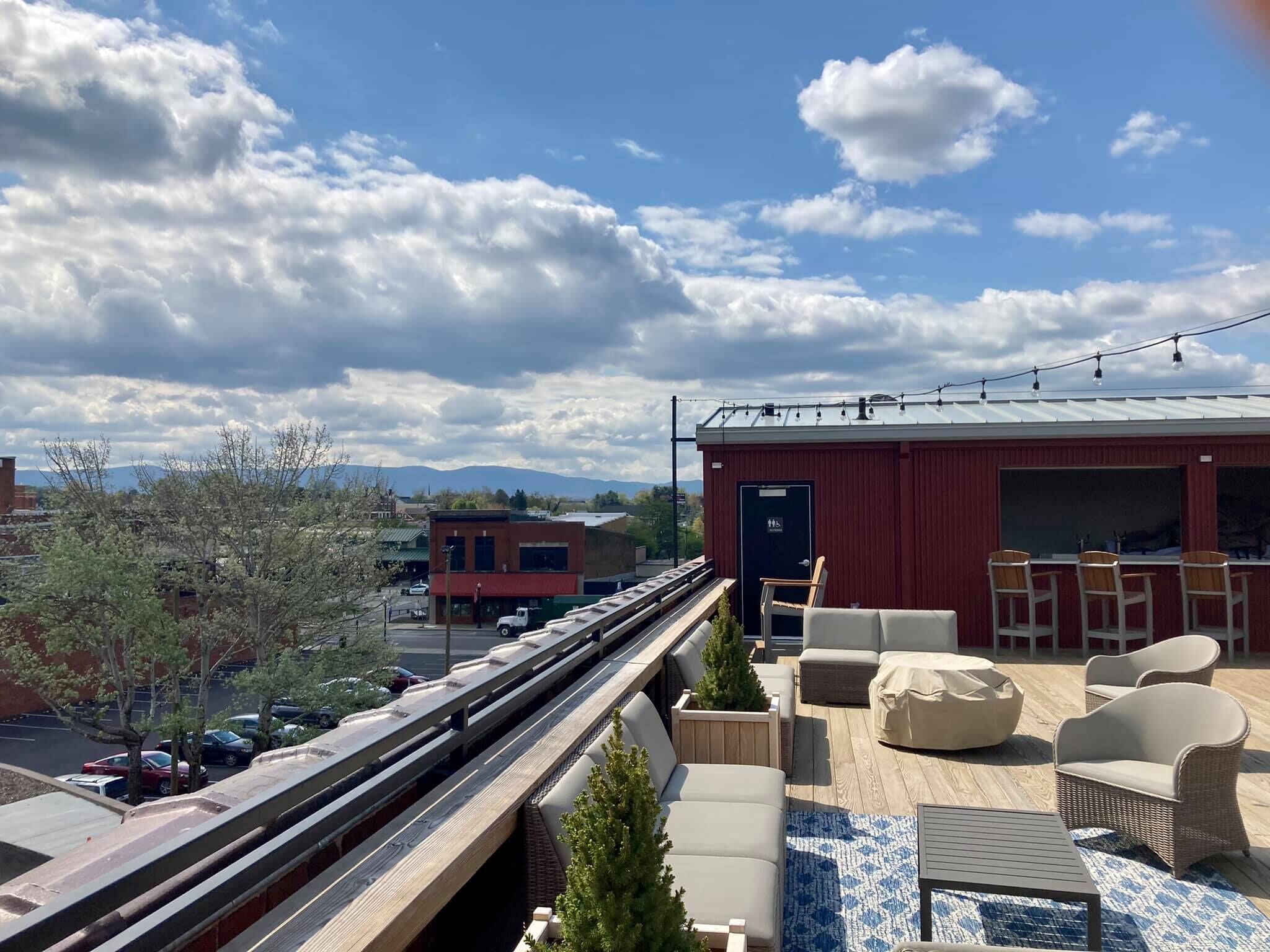 A rooftop terrace with outdoor seating, a red building, and a view of the city and mountains.