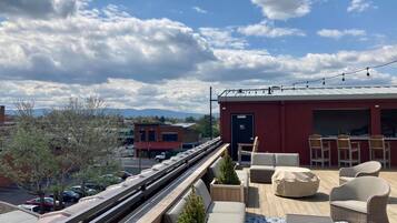 A rooftop terrace with outdoor seating, a red building, and a view of the city and mountains.