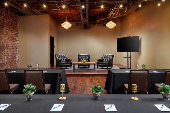 Un salón de conferencias con una larga mesa, sillas negras, un televisor y plantas decorativas.