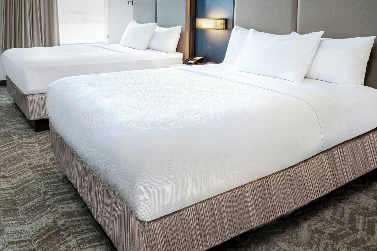 A hotel room with two beds, a carpeted floor, a nightstand with a lamp, and a window with curtains.