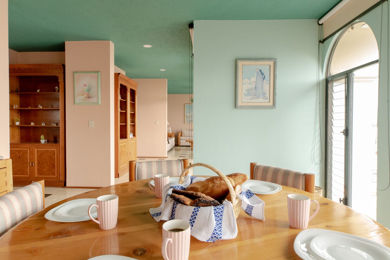 Ein Essbereich mit einem Holztisch, gedeckt für vier Personen, umgeben von gestreiften Stühlen. Es gibt zwei Holzschränke mit Glastüren, einen Korbwäscher mit Brot und ein gerahmtes Bild an der Wand.