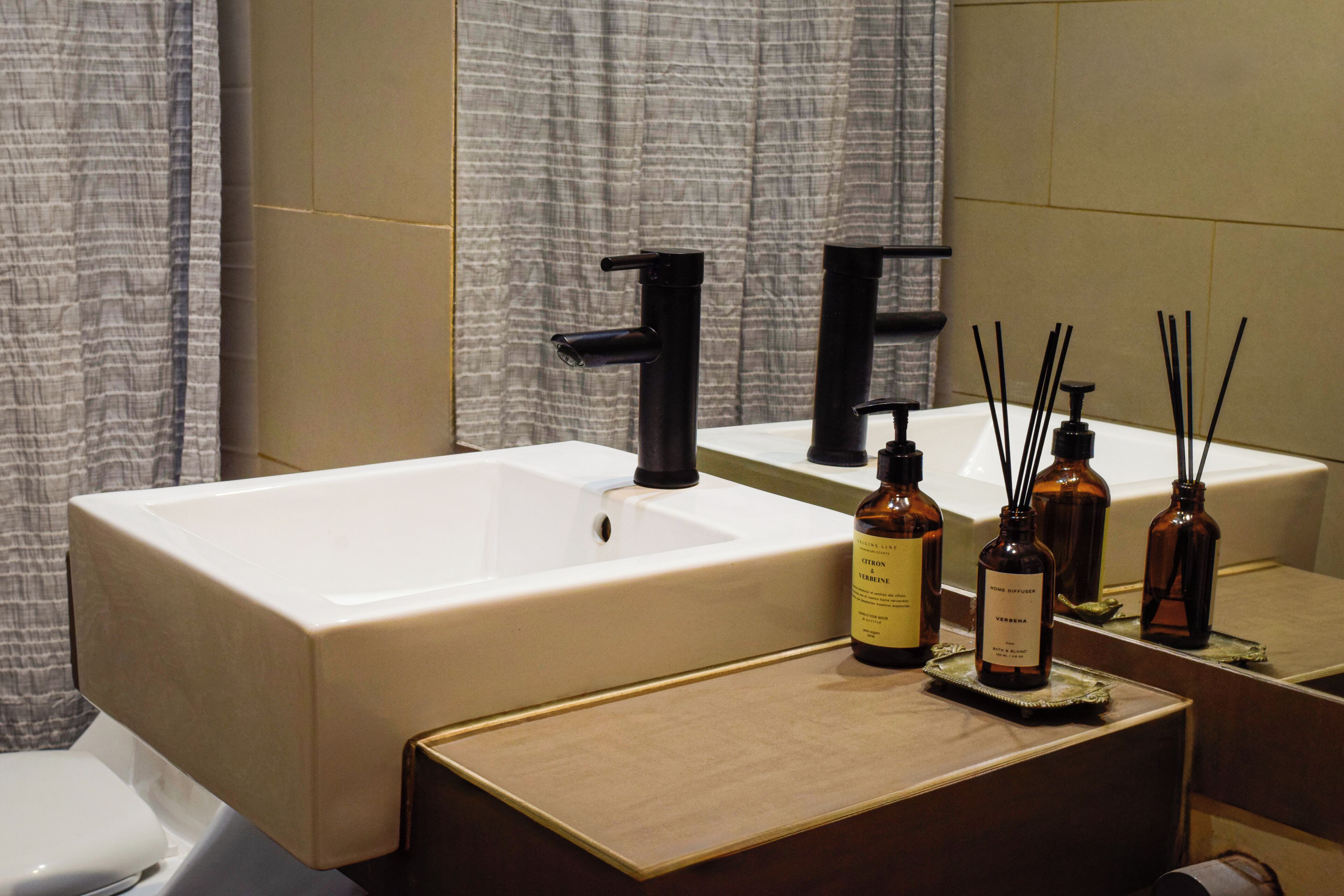 A bathroom with a large rectangular sink, two black faucets, and a mirror above it. There are three bottles on the sink's edge, and a toilet is visible to the left.