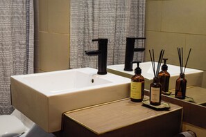 A bathroom with a large rectangular sink, two black faucets, and a mirror above it. There are three bottles on the sink's edge, and a toilet is visible to the left.
