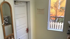 Una habitación con una puerta blanca, un ventanal con vista a un árbol, un espejo y un zapatero.