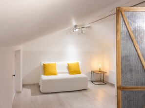 A minimalist room with a white sofa, two yellow cushions, a small table with a lamp, and a wooden-framed window. - Homerez - Cozy House in Cirey-sur-Vezouze (Cirey-sur-Vezouze)