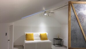 Una habitación pequeña con paredes blancas, una cama, dos almohadas amarillas, una mesita con una lámpara y un techo inclinado.