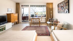 Una habitación de hotel con cama, televisor, mesa de comedor con sillas, sofá, un cuadro en la pared y vistas al exterior a través de la ventana.