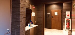 Habitación de hotel con puerta de madera, un lavamanos pequeño con un teléfono y una botella, y un extintor en la pared.