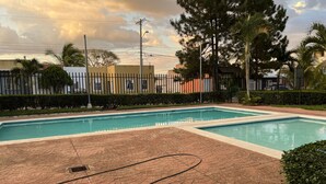Piscina retangular com um caminho de tijolos que a leva até a borda, cercada por uma grade e vegetação.
