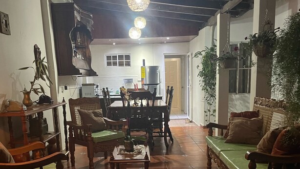 Uma sala de estar aconchegante com uma mesa de jantar, cadeiras e um sofá. Há uma área de cozinha ao fundo, com uma geladeira e um micro-ondas.
