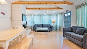 Een woonkamer met een houten eettafel, een grijze bank, een witte fauteuil, een blauw gestreept gordijn en een plafondventilator.