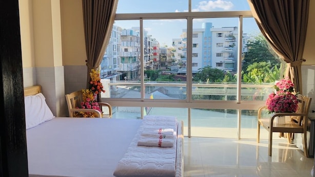 Habitación de hotel con una cama grande, vistas de la ciudad a través de una ventana y un balcón con sillas y flores.