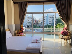 Une chambre d’hôtel avec un grand lit, une vue sur la ville par la fenêtre et un balcon agrémenté de chaises et de fleurs.