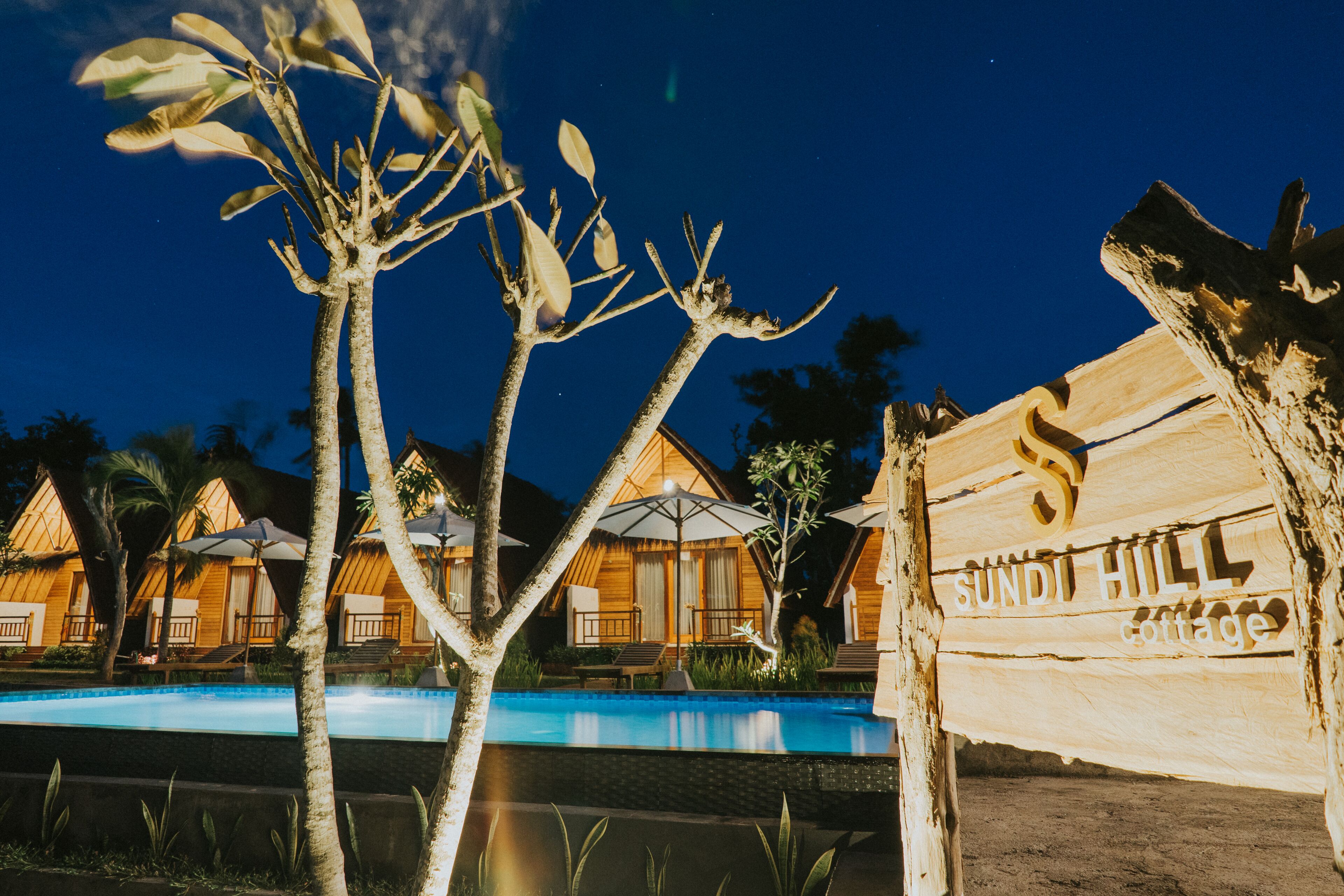 A wooden sign with "Sundi Hill Cottage" written on it, a swimming pool, and wooden cottages with umbrellas.