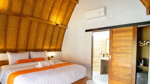 Un dormitorio con techo de madera, una cama con ropa de cama blanca y naranja, una mesita de noche y un baño visible a través de una puerta abierta.