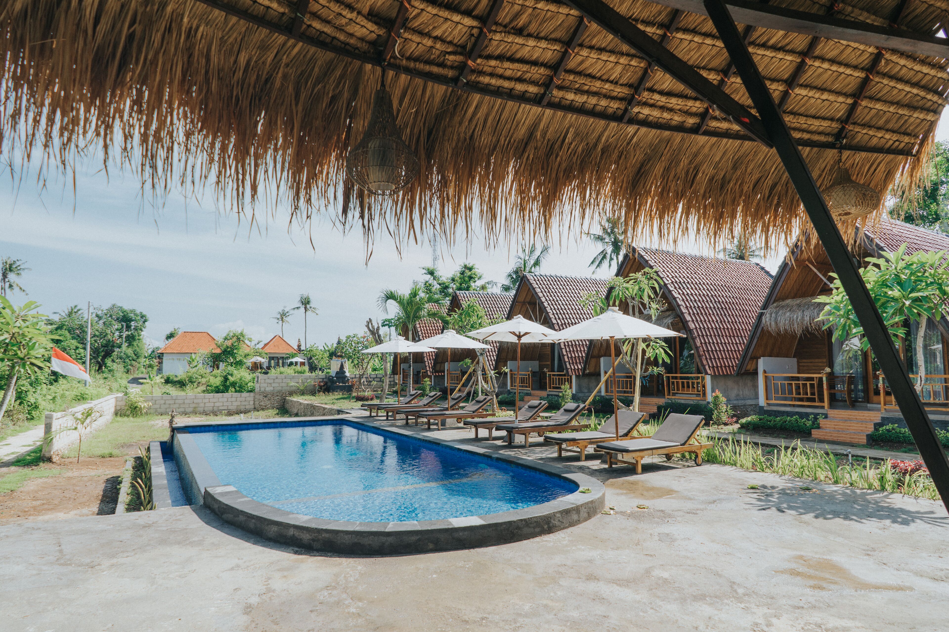 Un área de piscina con sillones de playa y sombrillas, rodeada de edificios con techo de paja y palmeras.