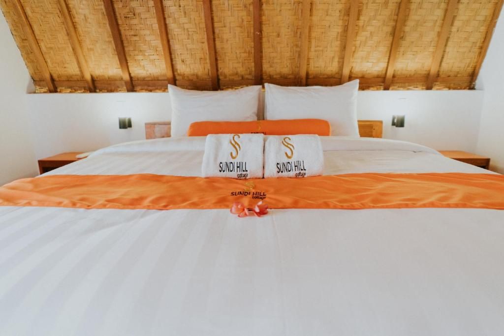 A bed with white linens and orange accents, two pillows with 'Sundhill' branding, and a woven ceiling.