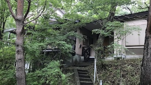 A residential building with a staircase leading to a covered entrance, surrounded by dense greenery. - Nasukogen Holiday (Nasu)