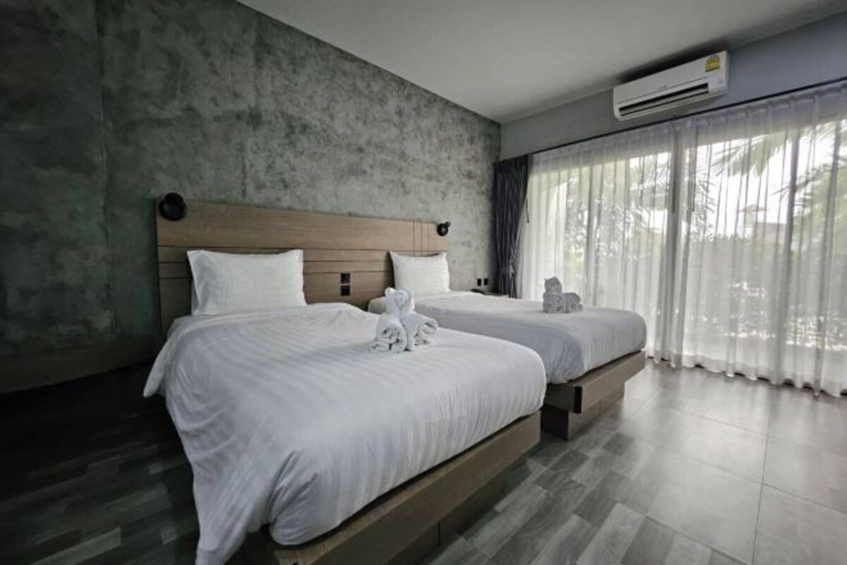 Habitación de hotel con dos camas, una pared de concreto, un equipo de aire acondicionado y ventanas amplias con cortinas.