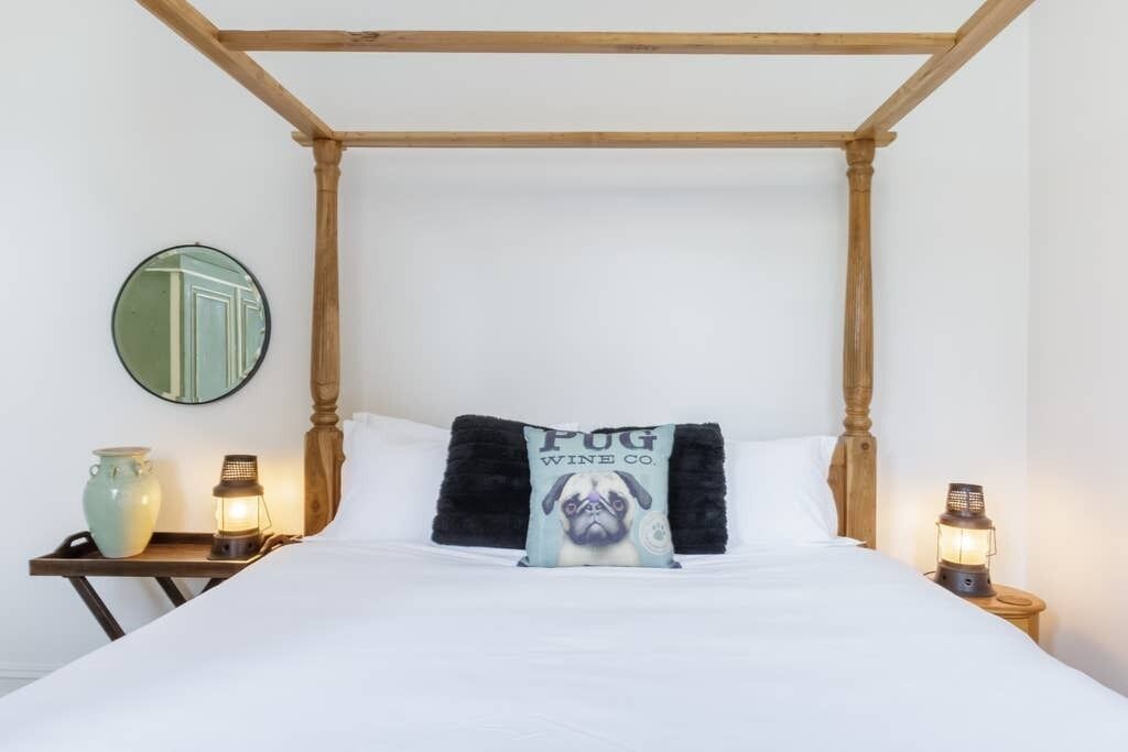 Un dormitorio con cama a dosel, un espejo redondo, una mesita de noche con un jarrón y un farol, y un cojín con diseño de pug.