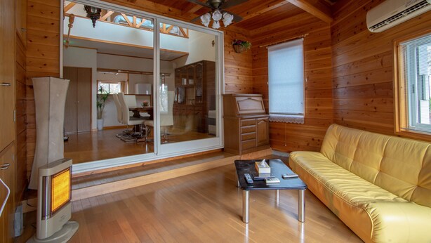 Interior de una cabaña de madera con sofá, mesa, calentador, ventilador de techo y ventana con persianas.