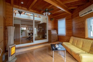 A wooden cabin interior with a sofa, a table, a heater, a ceiling fan, and a window with blinds.