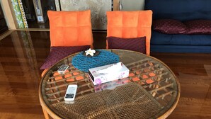 Una mesa redonda de vidrio con una base tejida, dos sillas con cojines anaranjados, un mantel tejido azul, una caja de pañuelos blancos y un teléfono inteligente.