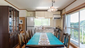 Ein Esszimmer mit einem langen Tisch, Stühlen, einem Holzschrank, einem Kühlschrank, einer Mikrowelle und einem Fenster mit Blick auf Grünflächen.