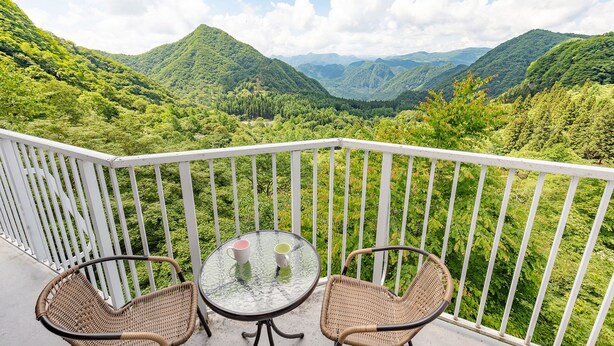 Ein Balkon mit Korbstühlen und einem Glastisch, mit Blick auf ein üppiges, grünes Tal mit Bergen in der Ferne.