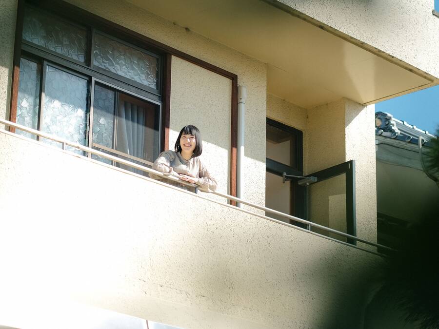 A person leaning on a balcony railing of a building with a textured facade and a window with a decorative pattern.