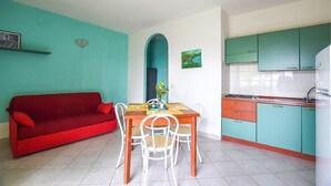 A compact living space with a red sofa, a dining table with chairs, and a kitchen area with teal cabinets and a white refrigerator.