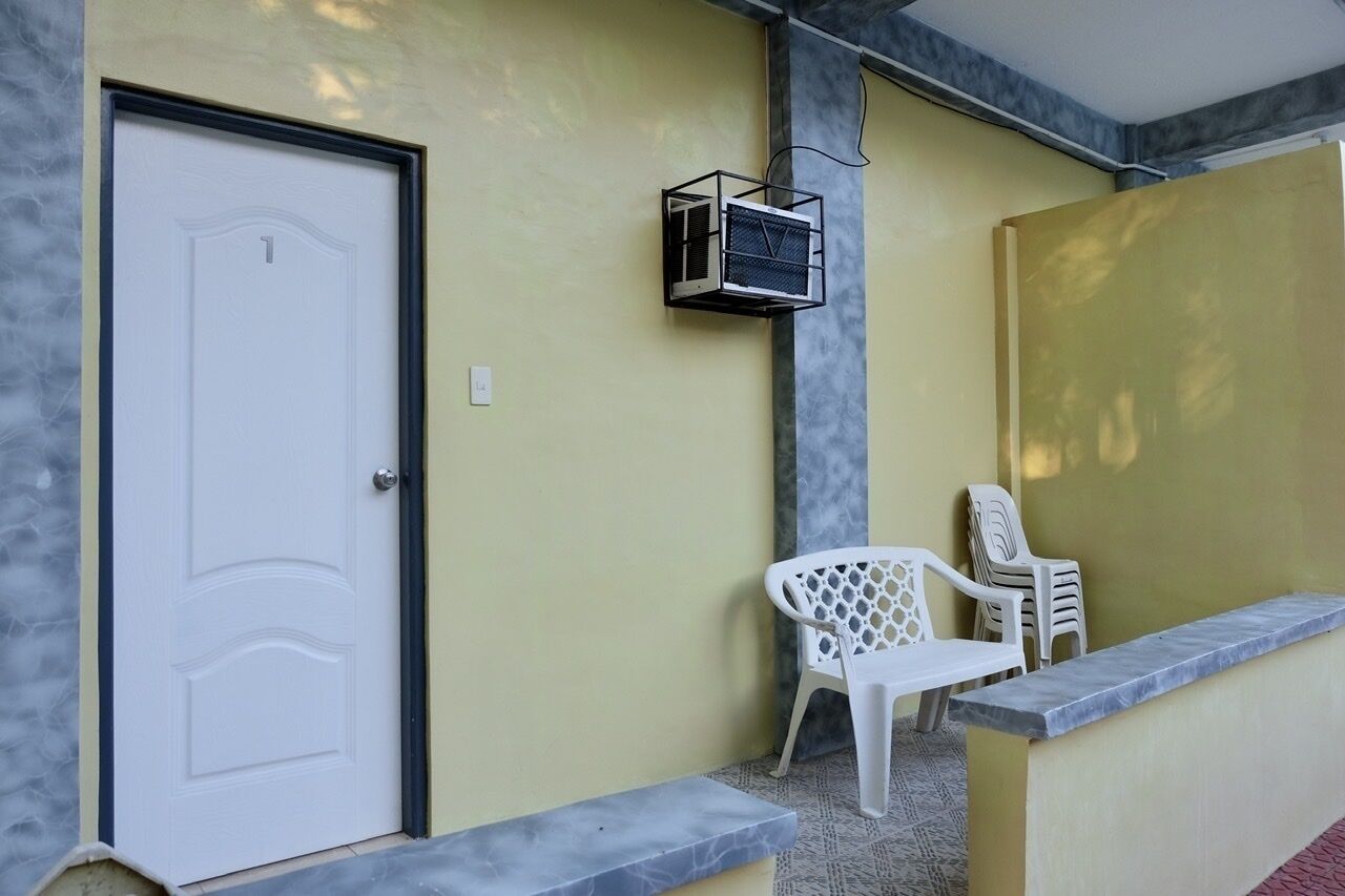 A room with a white door, a small air conditioning unit mounted on the wall, and two white plastic chairs against a yellow wall.