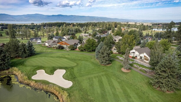 Un campo de golf con un estanque, casas y montañas al fondo.