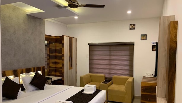 Quarto de hotel moderno com uma cama grande, ventoinha no teto, cabeceira de madeira, área de estar com sofá e poltronas, e uma televisão sobre um suporte de madeira.