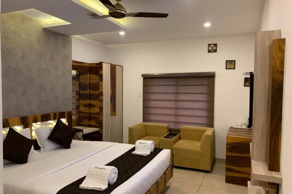 A modern hotel room with a large bed, a ceiling fan, a wooden headboard, a seating area with a sofa and armchairs, and a TV on a wooden stand.
