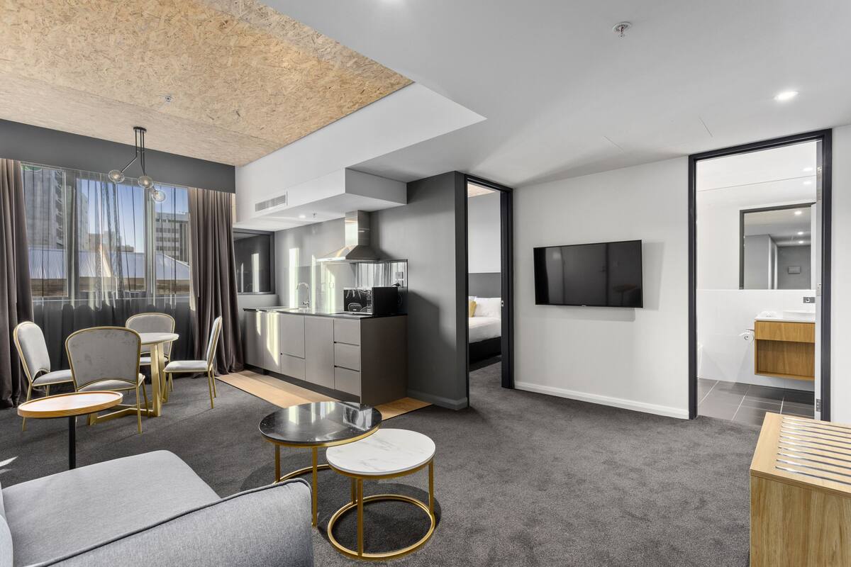A modern hotel room with a sofa, two round tables, a kitchen area, a TV, and a bathroom.