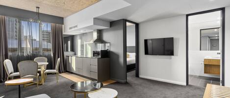 A modern hotel room with a sofa, two round tables, a kitchen area, a TV, and a bathroom.