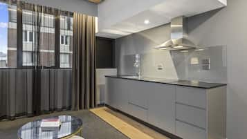 A modern kitchen with grey cabinets, a black countertop, and a stainless steel range hood. There is a window with curtains, a small round table with a book, and a wooden floor.