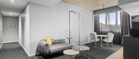 Une chambre d’hôtel moderne avec un canapé gris, une petite table ronde, un coin repas avec des chaises et un téléviseur posé sur un meuble.