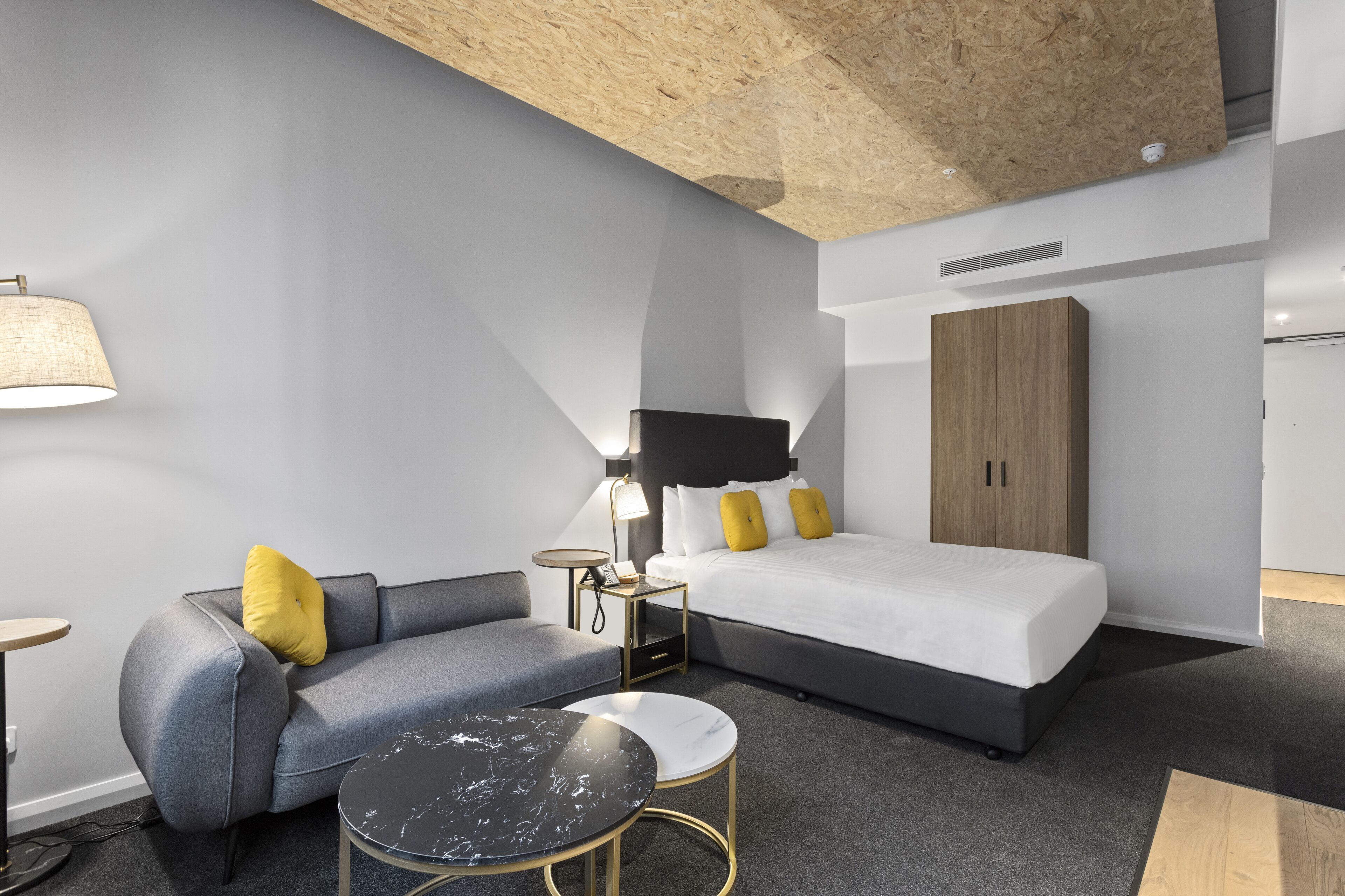 Een moderne hotelkamer met een grijze bank, een ronde salontafel, een bed met wit beddengoed en een houten garderobe.
