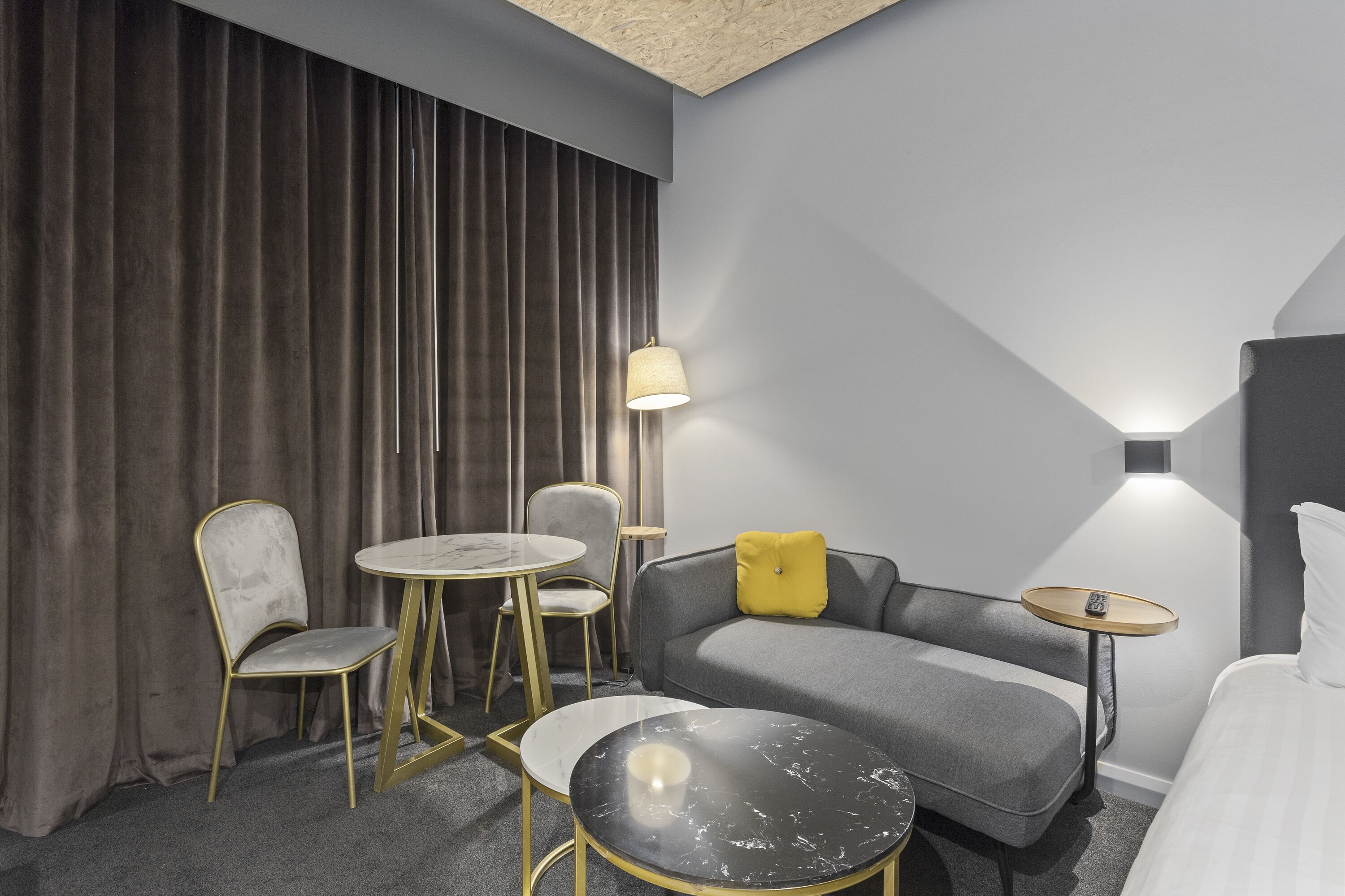 Ein modernes Hotelzimmer mit grauem Sofa, einem runden Couchtisch, einem kleinen Tisch mit Lampe, einem Sessel und einem Bett.