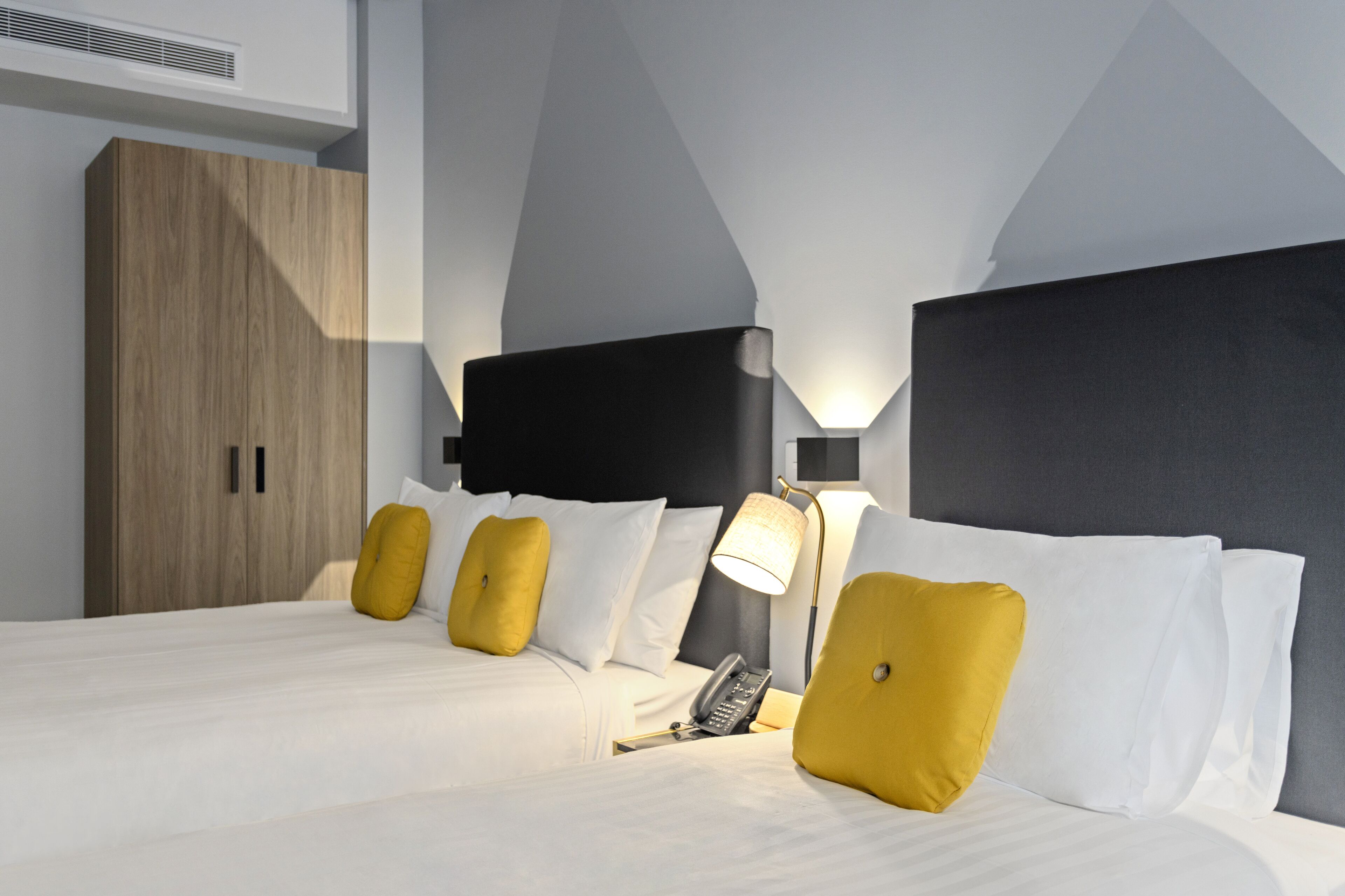 A hotel room with a modern design, featuring a large bed with white bedding and two yellow accent pillows, a bedside telephone, and a lamp on the nightstand.
